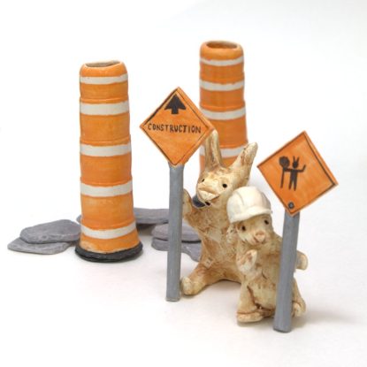 Deux figurines de lapins en céramique sont sur un site de construction. Ils tiennent des affiches signalétiques devant des cônes oranges et une pile de roches.